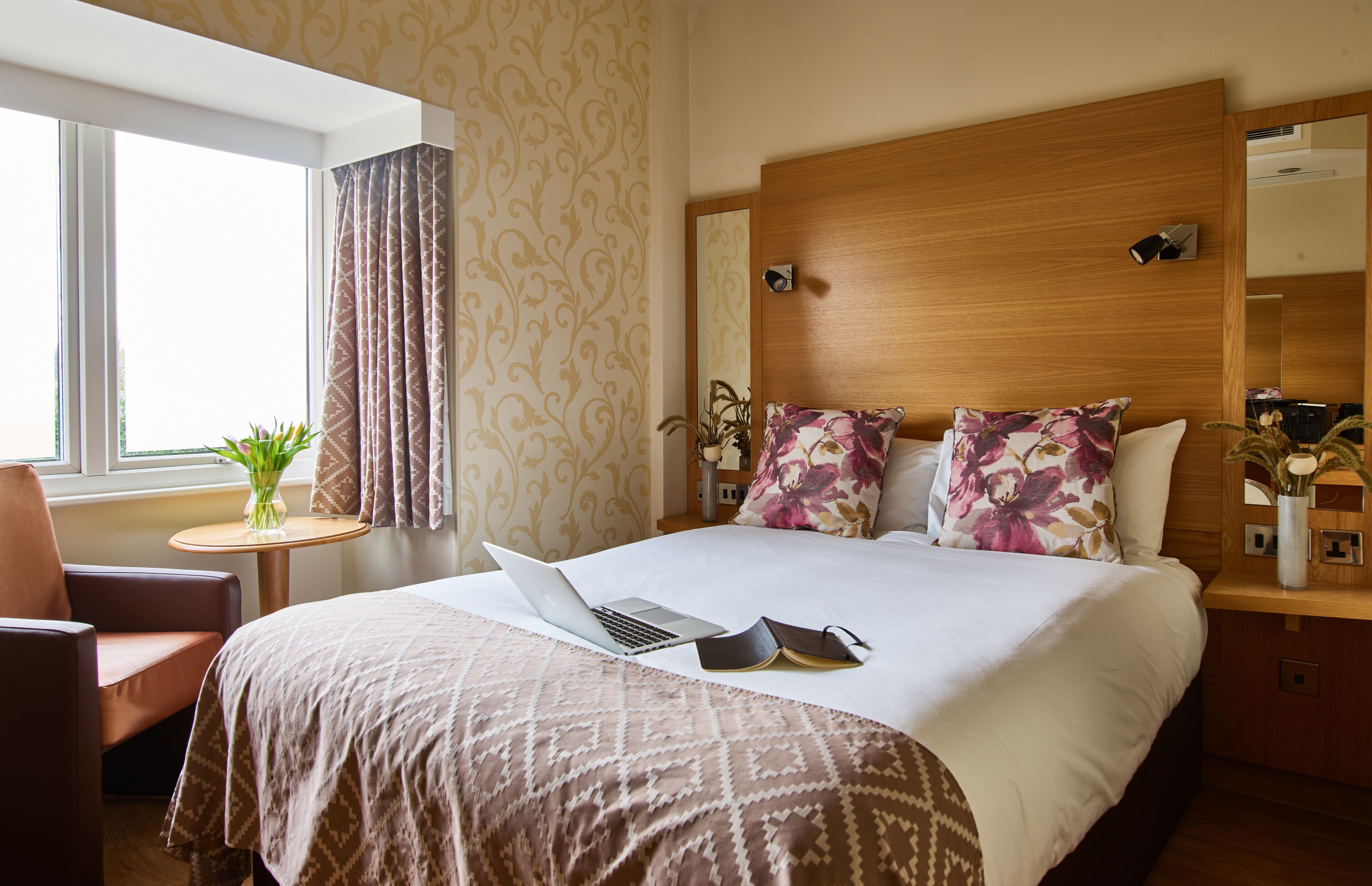 A hotel room with a large bed, a laptop, a book, and a vase with flowers on the nightstand.