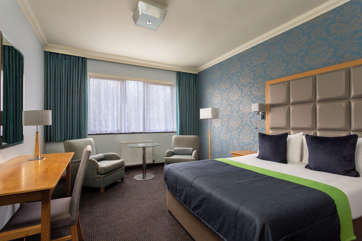 A hotel room with a large bed, two armchairs, a small table, and a window with curtains.