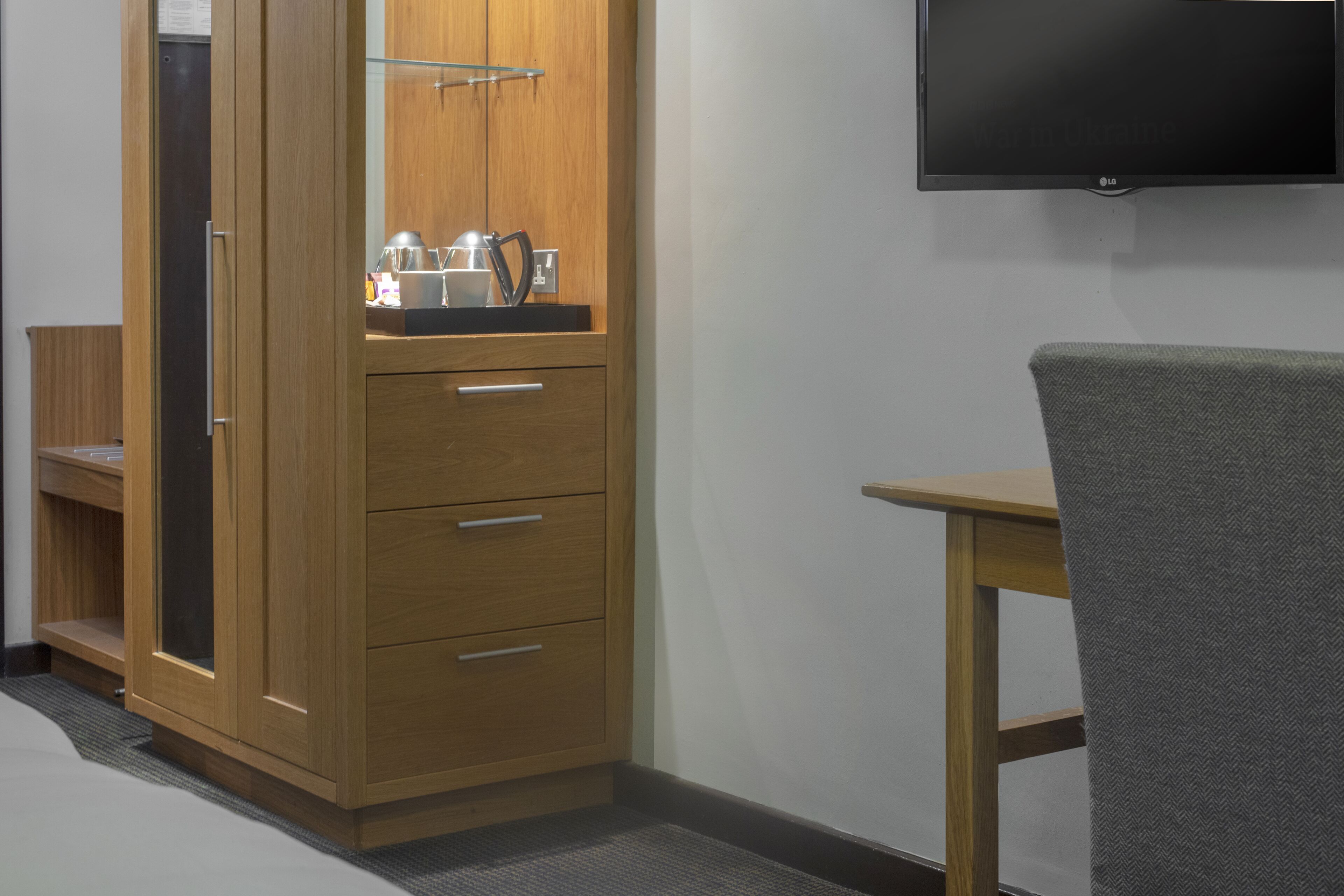 A wooden wardrobe with a mirror, a flat-screen TV mounted on the wall, and a small wooden table with a chair.