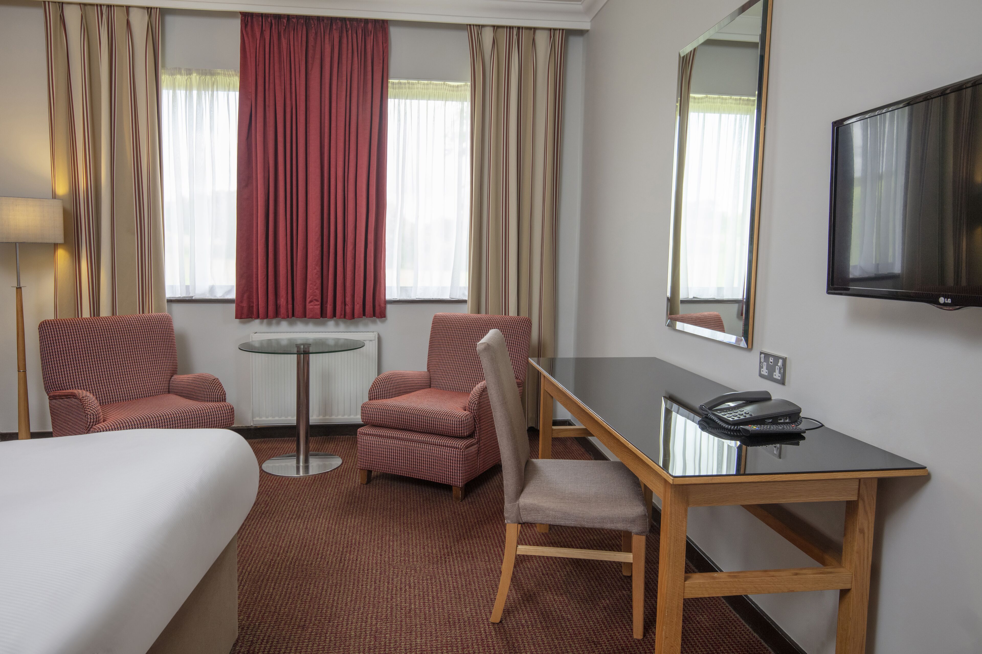 Habitación de hotel con cama, dos sillones, una mesita, un escritorio con teléfono y televisor en la pared.