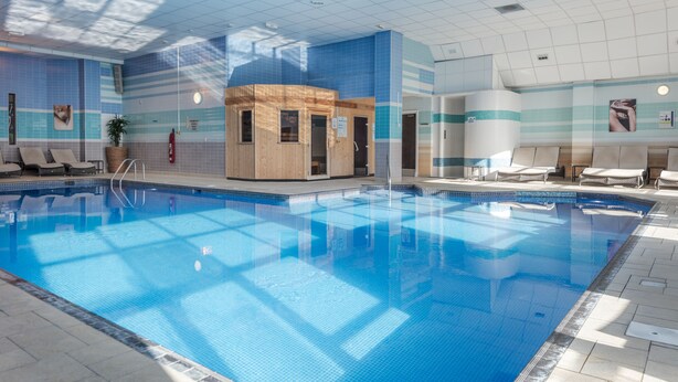Una piscina interior con agua azul cristalina, saunas de madera y sillones de descanso.