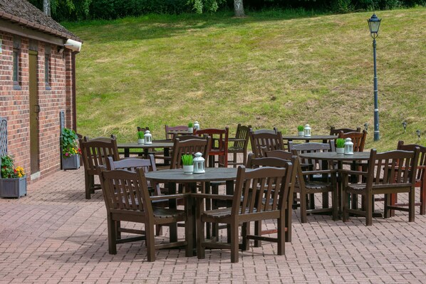 An outdoor patio with multiple wooden tables and chairs, surrounded by brick flooring, and a brick building with a window and door.