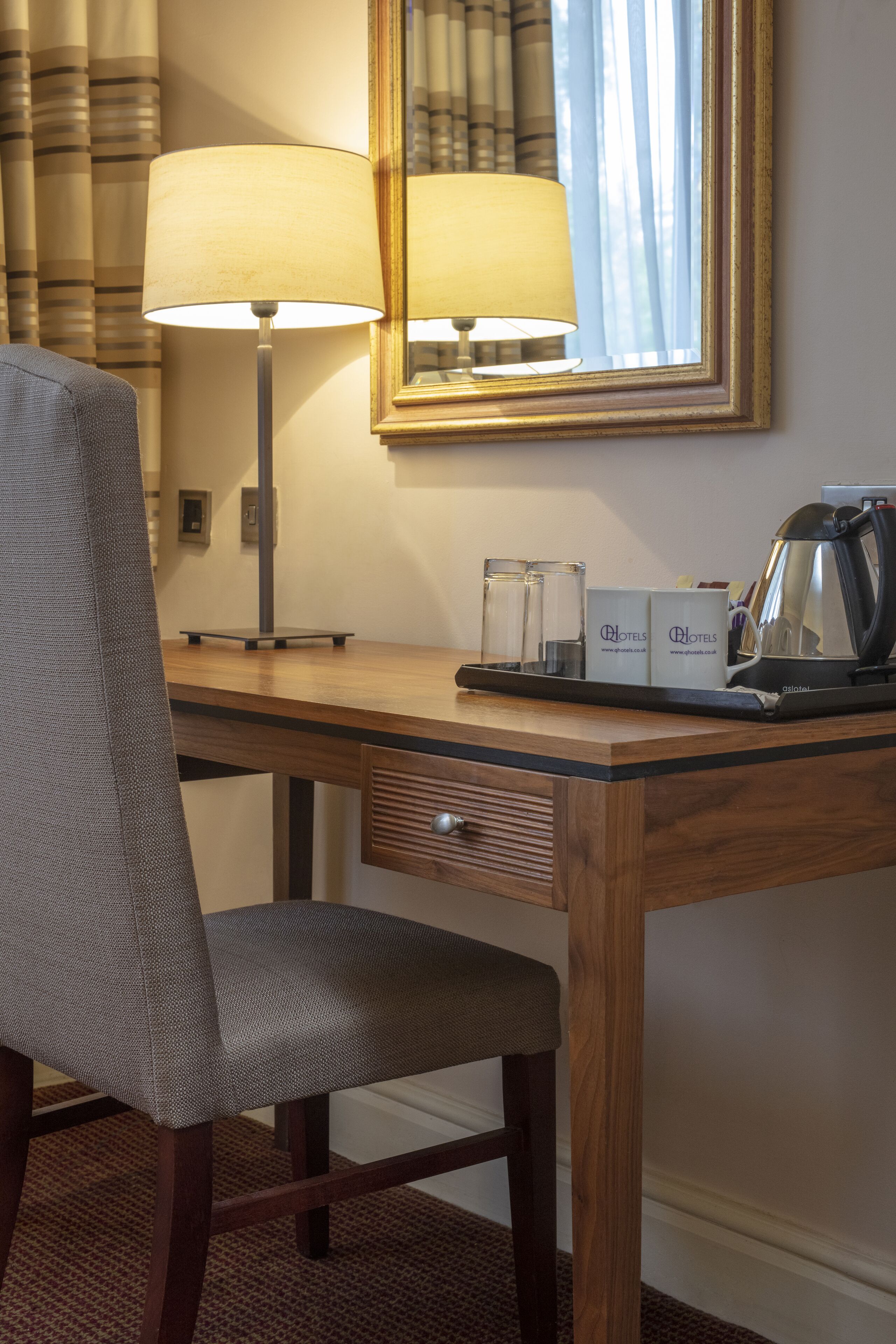 A hotel room desk with a lamp, a chair, a tray with glasses and a kettle, and a mirror on the wall.