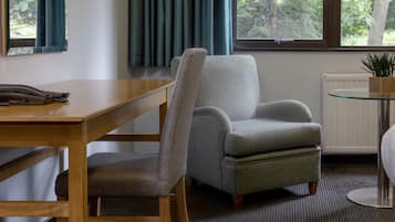 A hotel room with a wooden desk, a grey armchair, a small glass table, a radiator, and a window with curtains.