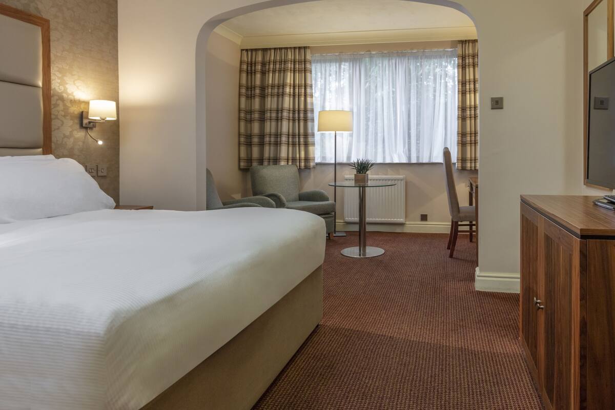 Ein Hotelzimmer mit einem großen Bett, einem Schreibtisch, einem Sessel, einem kleinen Tisch mit einer Lampe und einem Fernseher.