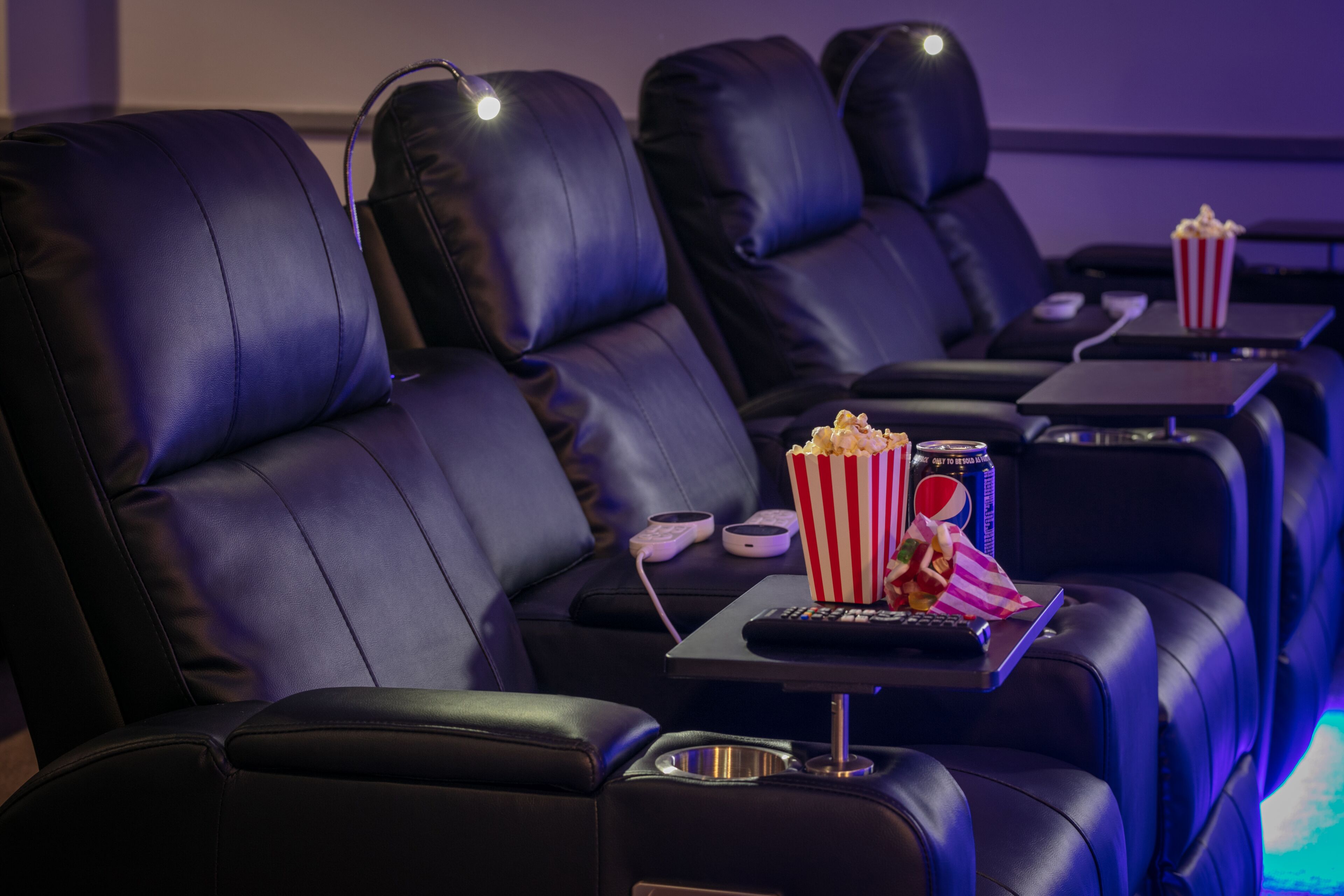 Filas de asientos de cuero negro en un cine, con reposabrazos individuales, cada uno con una mesita que sostiene un balde de palomitas y un control remoto.