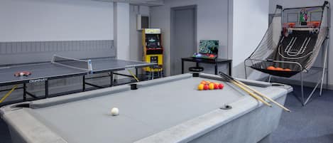 Una mesa de billar junto a una mesa de ping pong y un aro de baloncesto pequeño en una habitación con paredes blancas y una puerta.