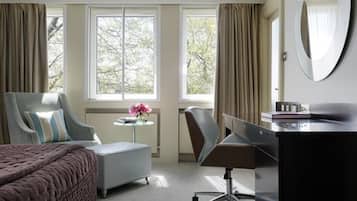 A hotel room with a bed, a desk, a chair, a small table with a vase of flowers, and large windows with curtains.