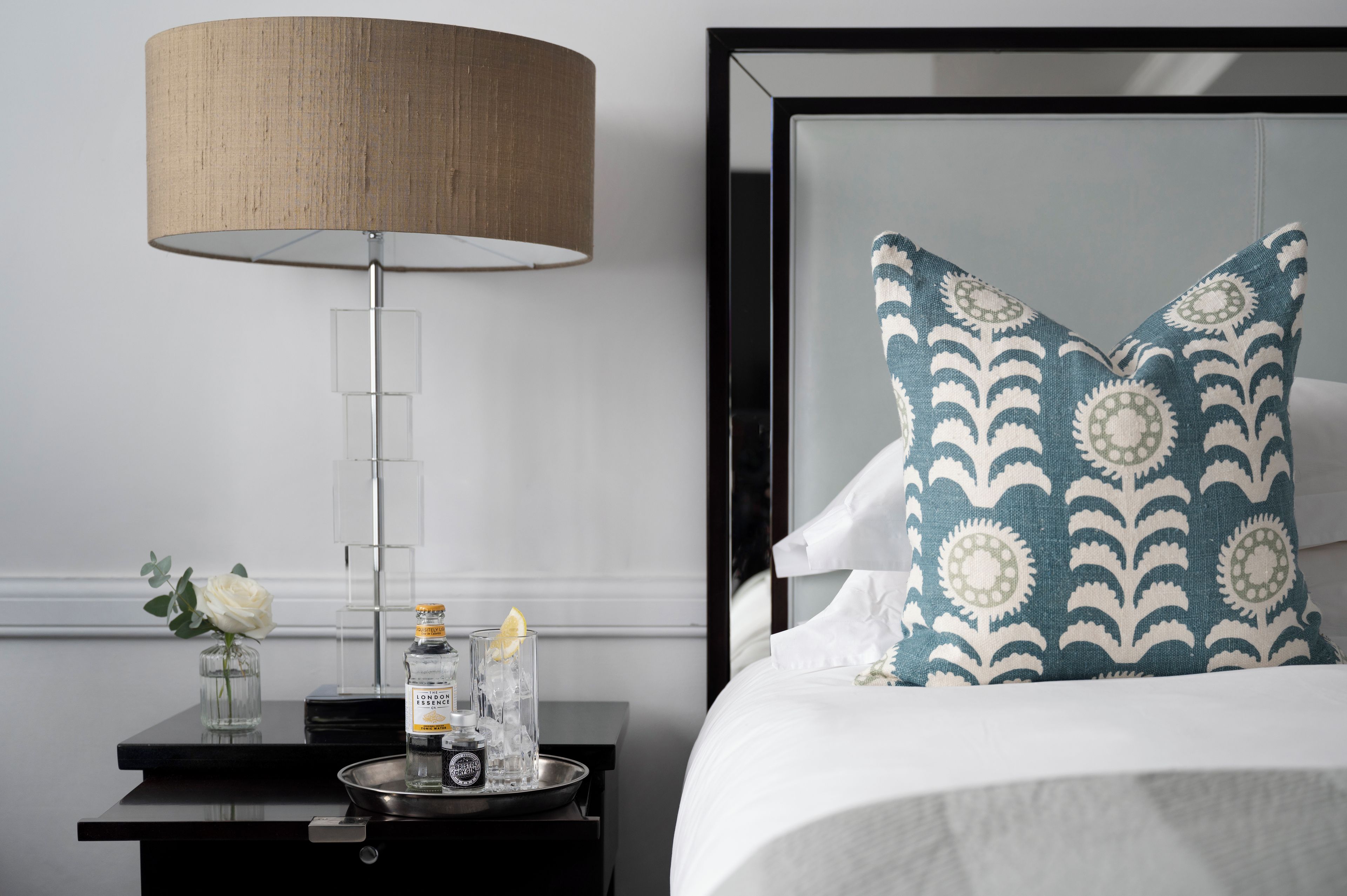 A neatly arranged hotel room with a bed, a bedside table with a lamp, a vase with a flower, and a tray with bottles.