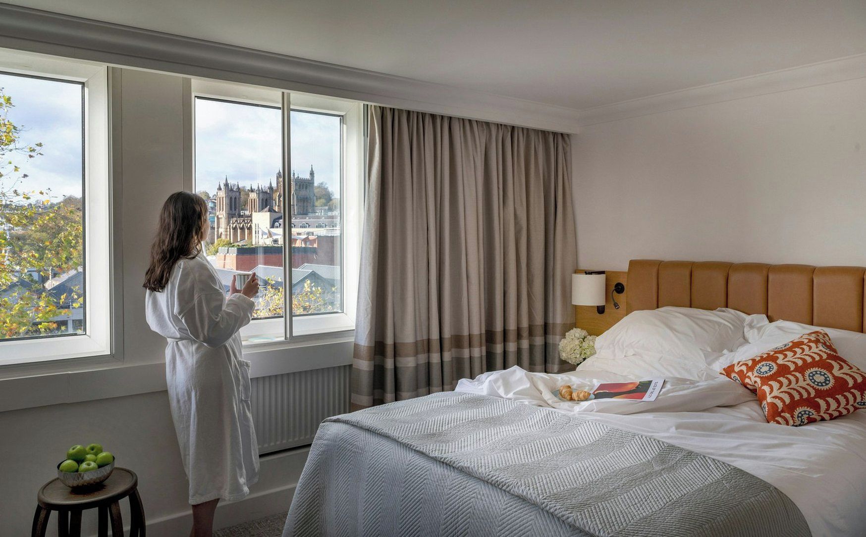 A hotel room with a large window, a bed with white linens, a headboard, and a bedside table with a bowl of apples.