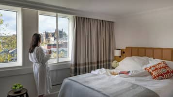 A hotel room with a large window, a bed with white linens, a headboard, and a bedside table with a bowl of apples.