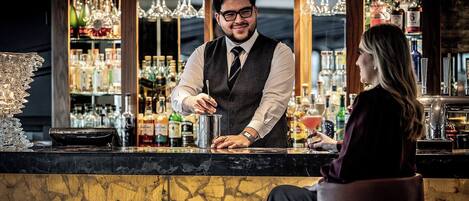 A bar area with a bartender preparing a drink, a customer seated, and a well-stocked bar with various bottles and a decorative glass structure.