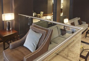 A hotel room with a seating area featuring a brown armchair with a white pillow, a glass partition, and a small table with a lamp.