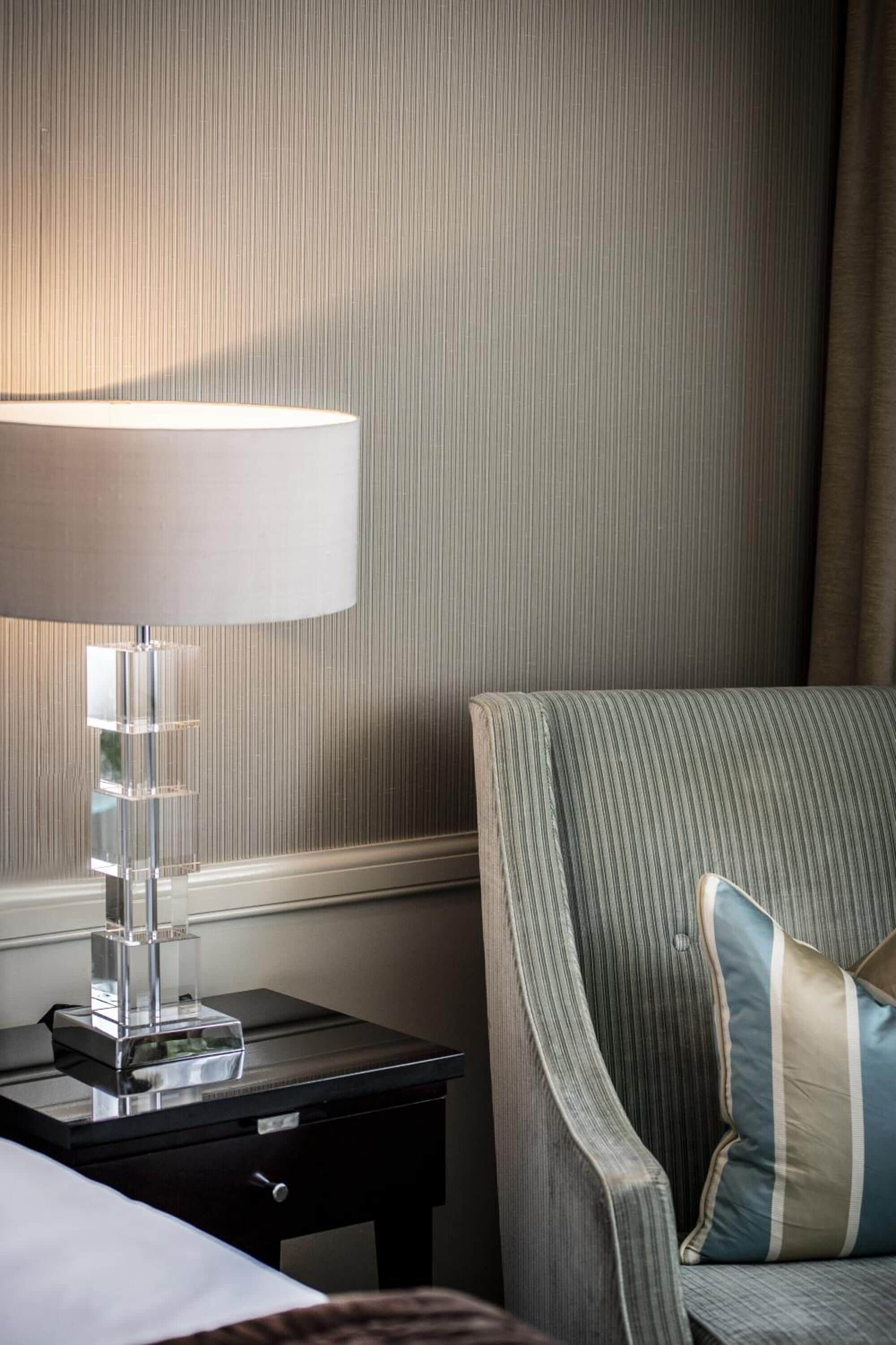 A hotel room with a striped armchair, a bedside table with a lamp, and a nightstand.
