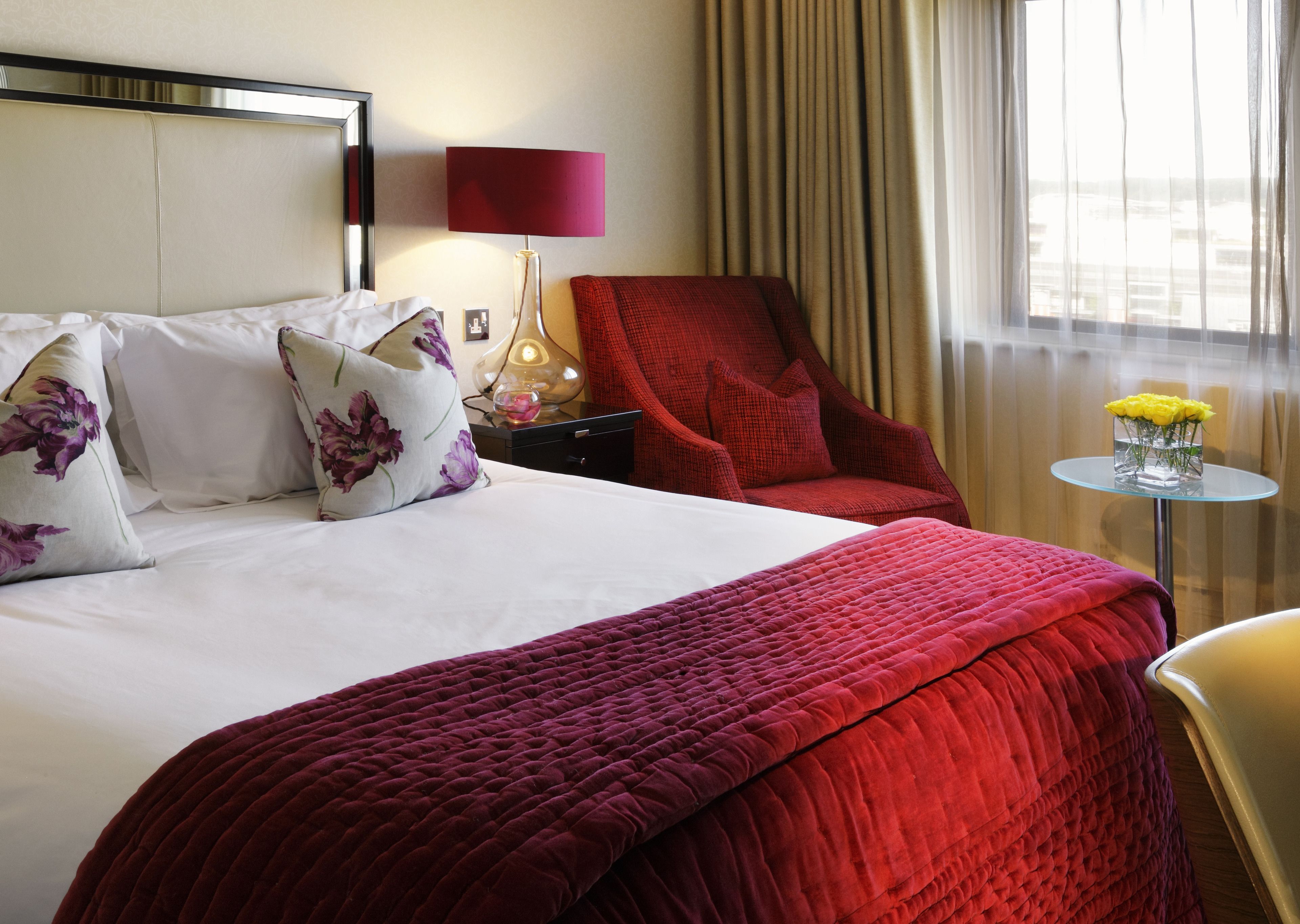 Une chambre d’hôtel avec un grand lit, un fauteuil rouge, une petite table sur laquelle est posé un vase de fleurs et une lampe sur une table de chevet.