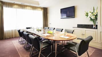 A conference room with a long table, black chairs, a flat-screen TV on the wall, and a vase with flowers on a counter.