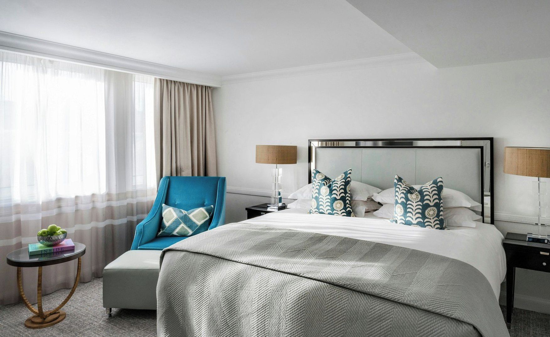 A hotel room with a bed, a chair, a nightstand, a lamp, and a small table with a bowl of fruit.