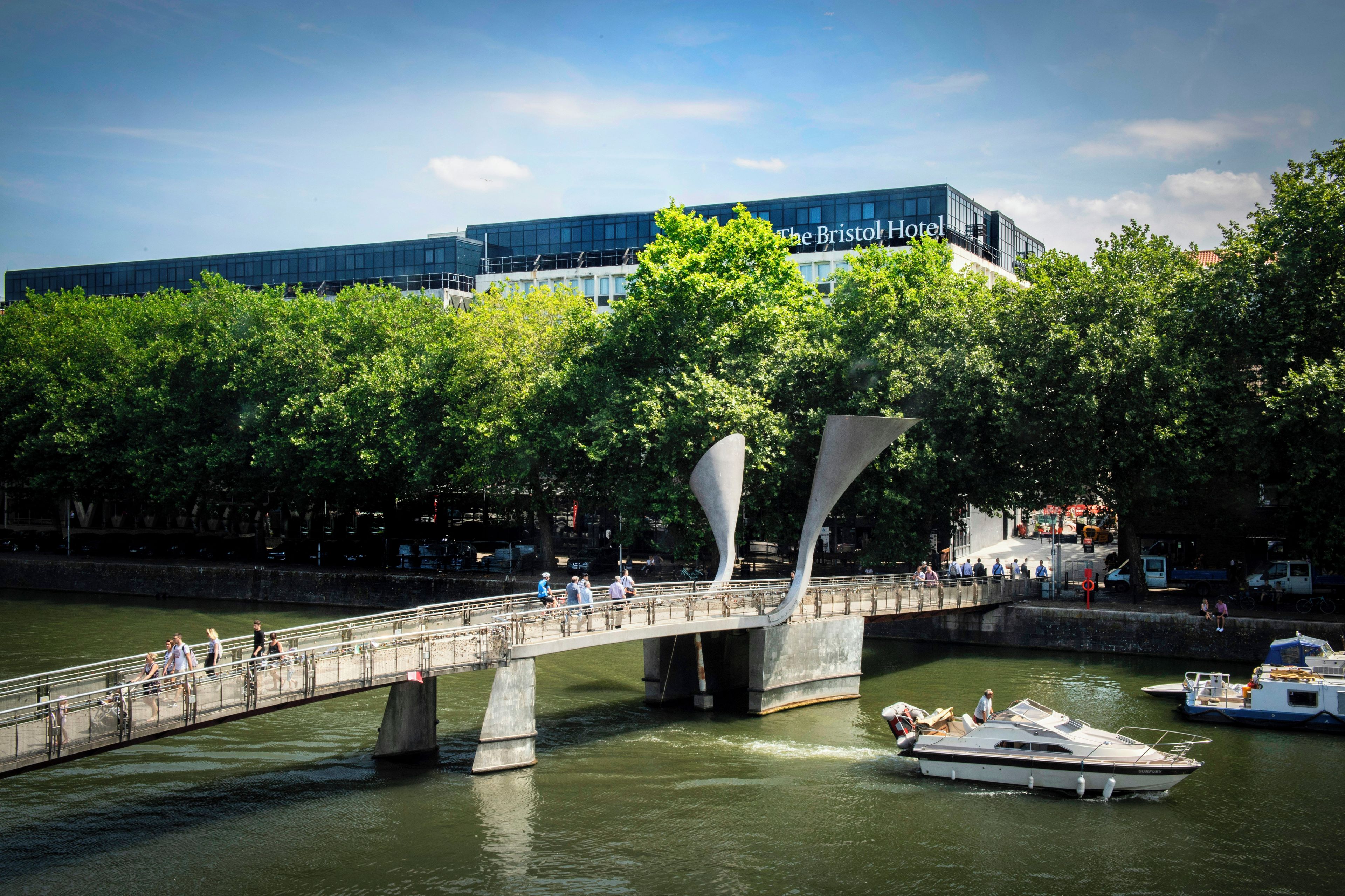 Un puente moderno con un diseño único, un río con barcos y un edificio con el cartel 'The Bristol Hotel' al fondo.