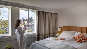 Hotelkamer met een groot raam, een bed met wit beddengoed, een houten hoofdeinde en een nachtkastje met een schaal appels.