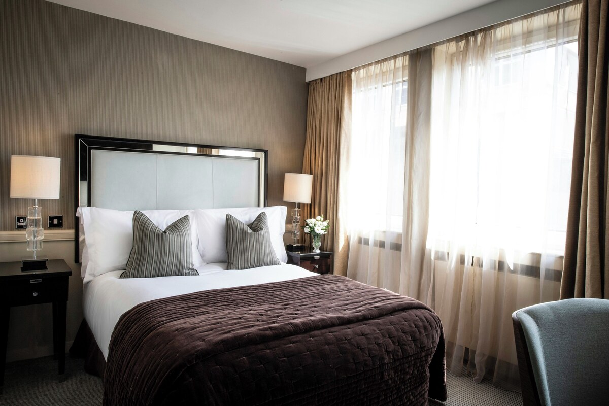 A hotel room with a large bed, two bedside tables, a chair, and a window with curtains.