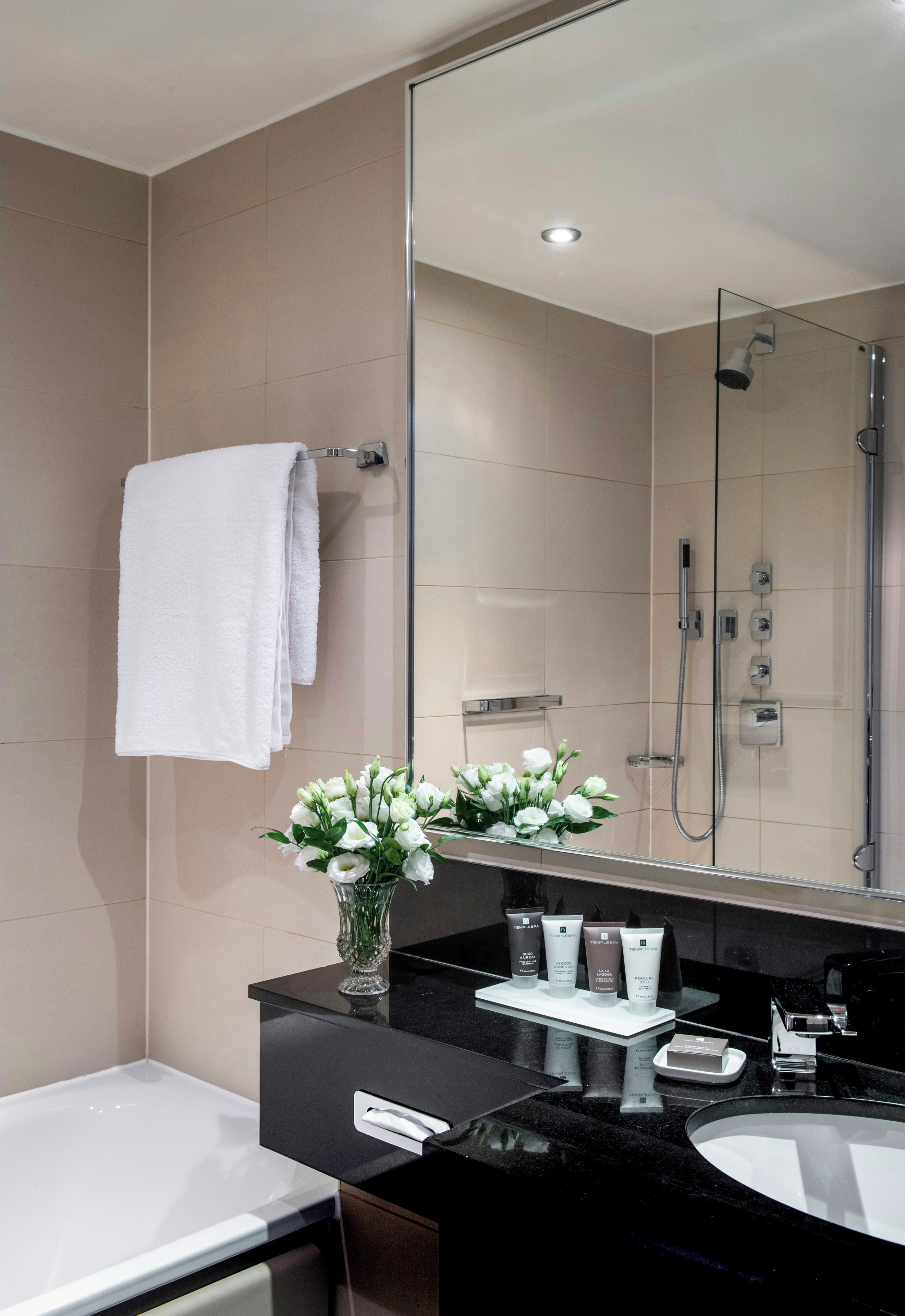 A modern bathroom with a large mirror, a white towel, a vase of flowers, and toiletries on the counter.