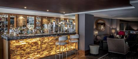 Zona de bar con mostrador de mármol, varios taburetes y una variedad de botellas y vasos.