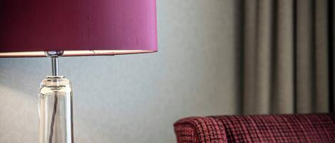 A clear glass table lamp with a metallic base and a dark-colored lampshade, positioned next to a dark-colored upholstered chair with a textured fabric.