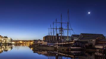 Um navio histórico atracado em um porto, com edifícios e um corpo d'água calma refletindo a paisagem.