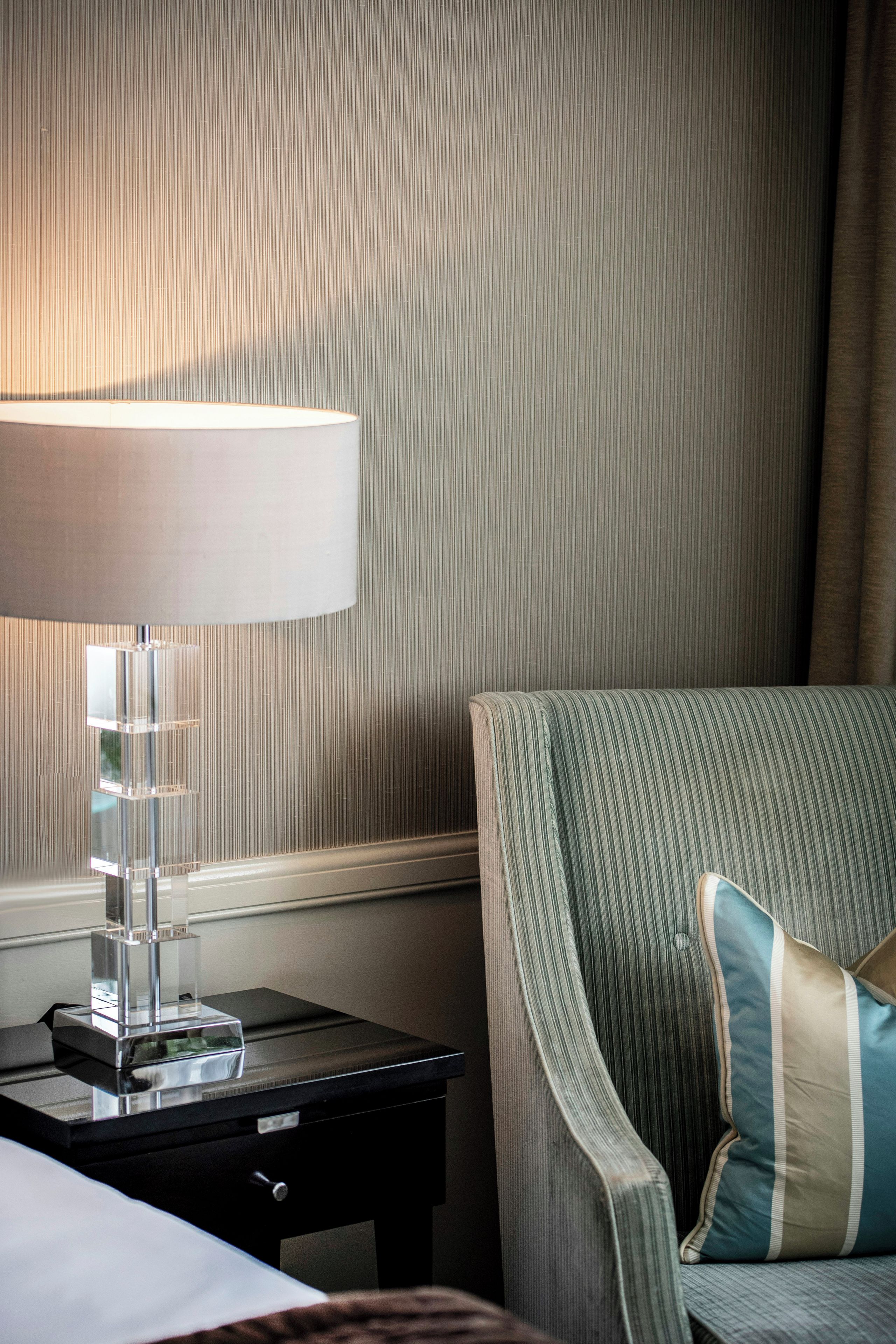 Una camera d'albergo con un letto, un comodino con una lampada, una sedia con un cuscino e una parete a righe.
