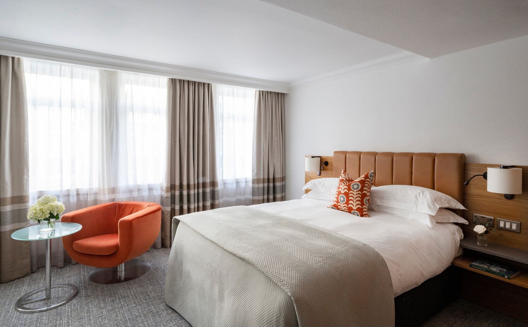 Una habitación de hotel con una cama grande, cabecera de madera, mesita de noche con lámpara, una mesa de vidrio con una silla y una ventana con cortinas.