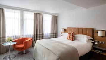 Una habitación de hotel con una cama grande, cabecera de madera, mesita de noche con lámpara, una mesa de vidrio con una silla y una ventana con cortinas.