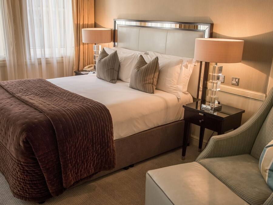 A hotel room with a large bed, two bedside lamps, a bedside table with a lamp, a chair, and a bedside table with a mirror.