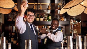 Twee bartenders in een bar, met flessen drank op de planken en een toog met diverse barbenodigdheden.