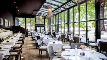 Un restaurant avec de longues tables et des chaises, de grandes fenêtres et des luminaires suspendus.