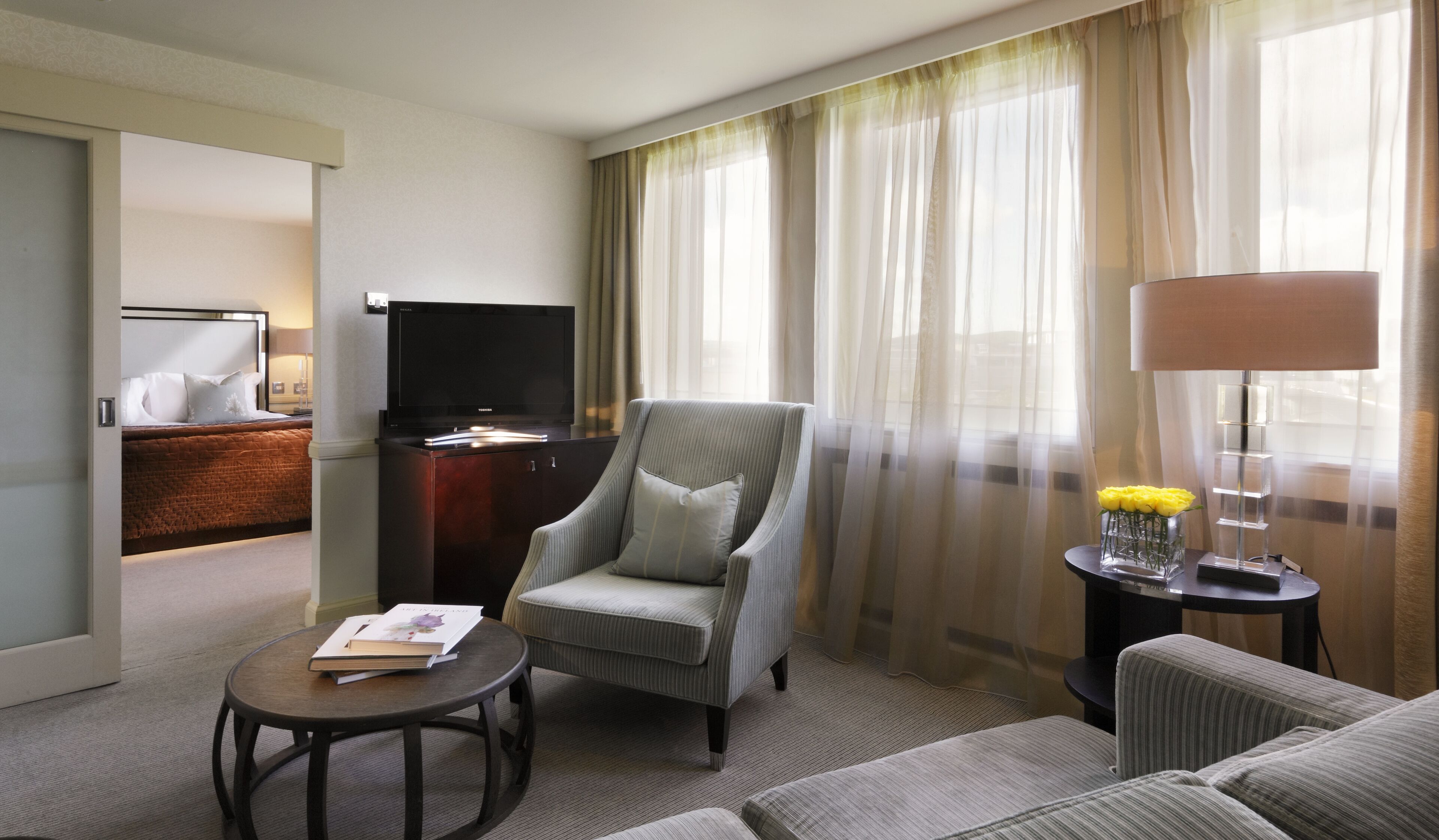 A hotel room with a bed, a TV, a chair, a sofa, a small table with a book, a lamp, and a vase with flowers.
