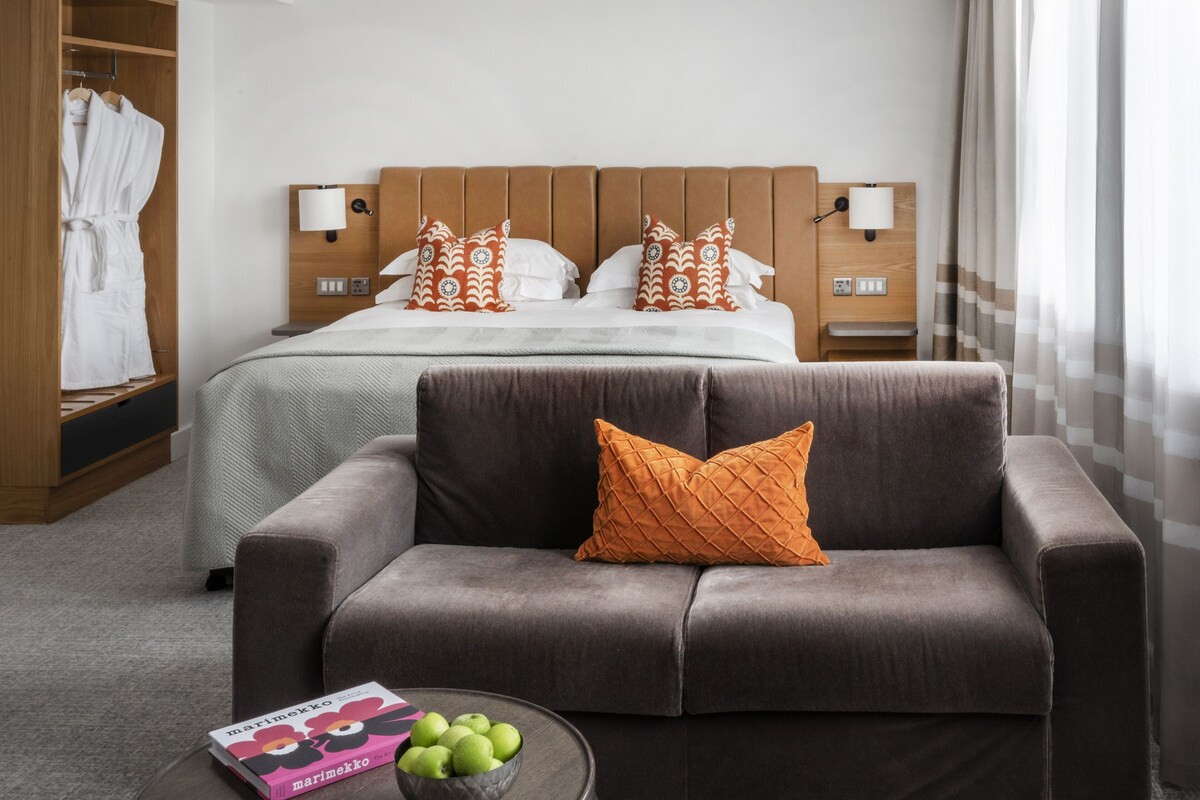 A hotel room with a bed, a sofa, a nightstand, a lamp, a wardrobe, and a bowl of apples on a table.