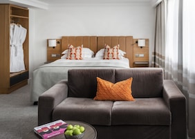 A hotel room with a bed, a sofa, a nightstand, a lamp, a wardrobe, and a bowl of apples on a table.