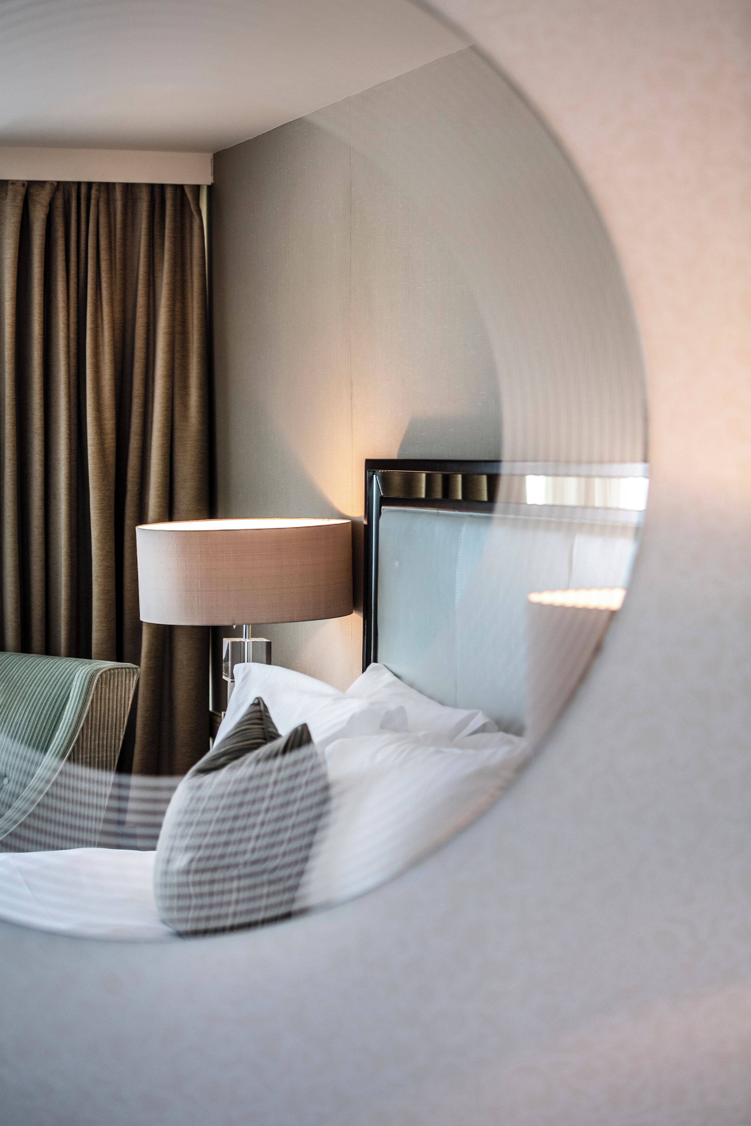 Quarto de hotel com cama, mesa de cabeceira, luminária e vista refletida em espelho circular.