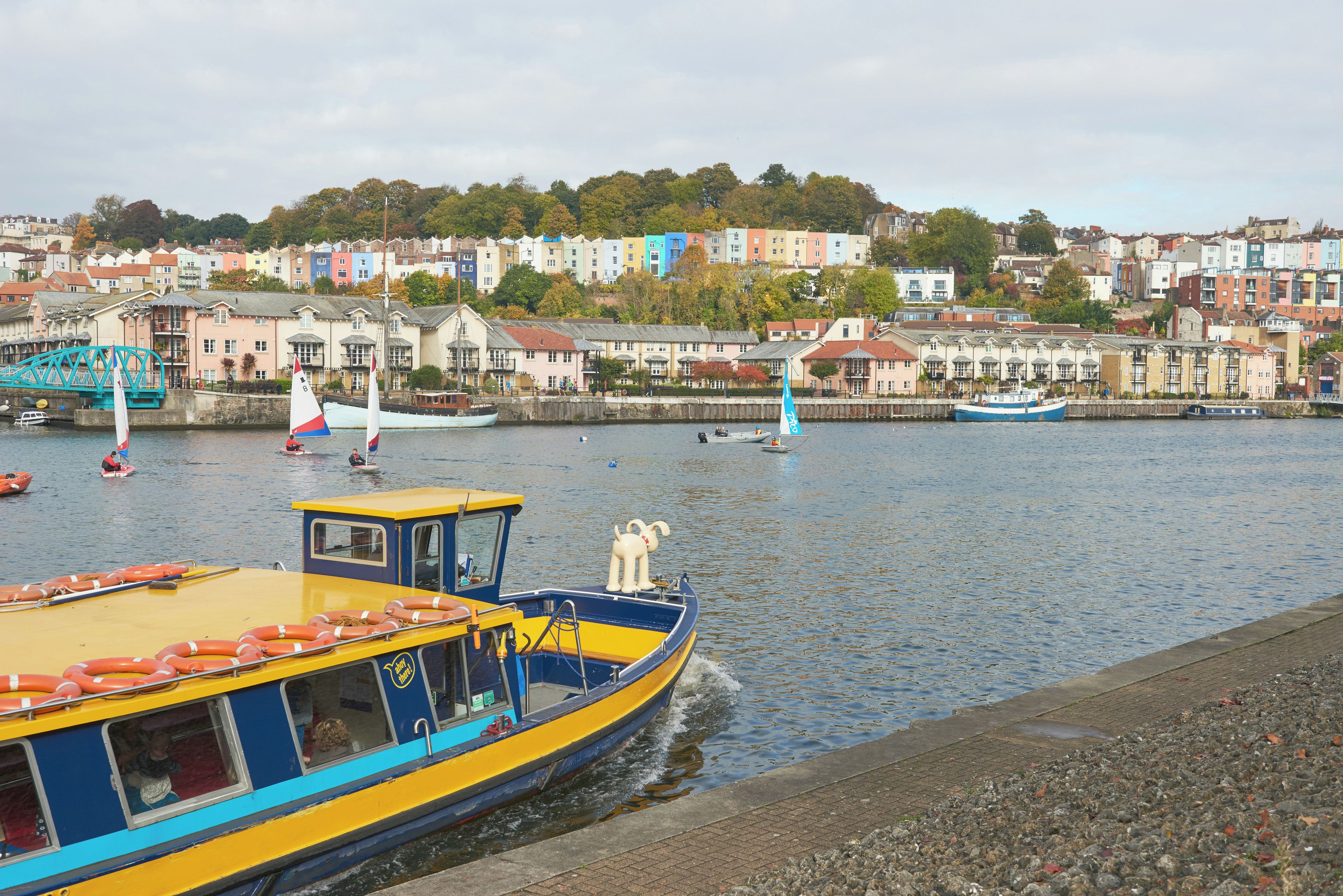 Une promenade en bord de mer bordée de bâtiments aux couleurs vives, un bateau jaune avec des bouées de sauvetage et un voilier.