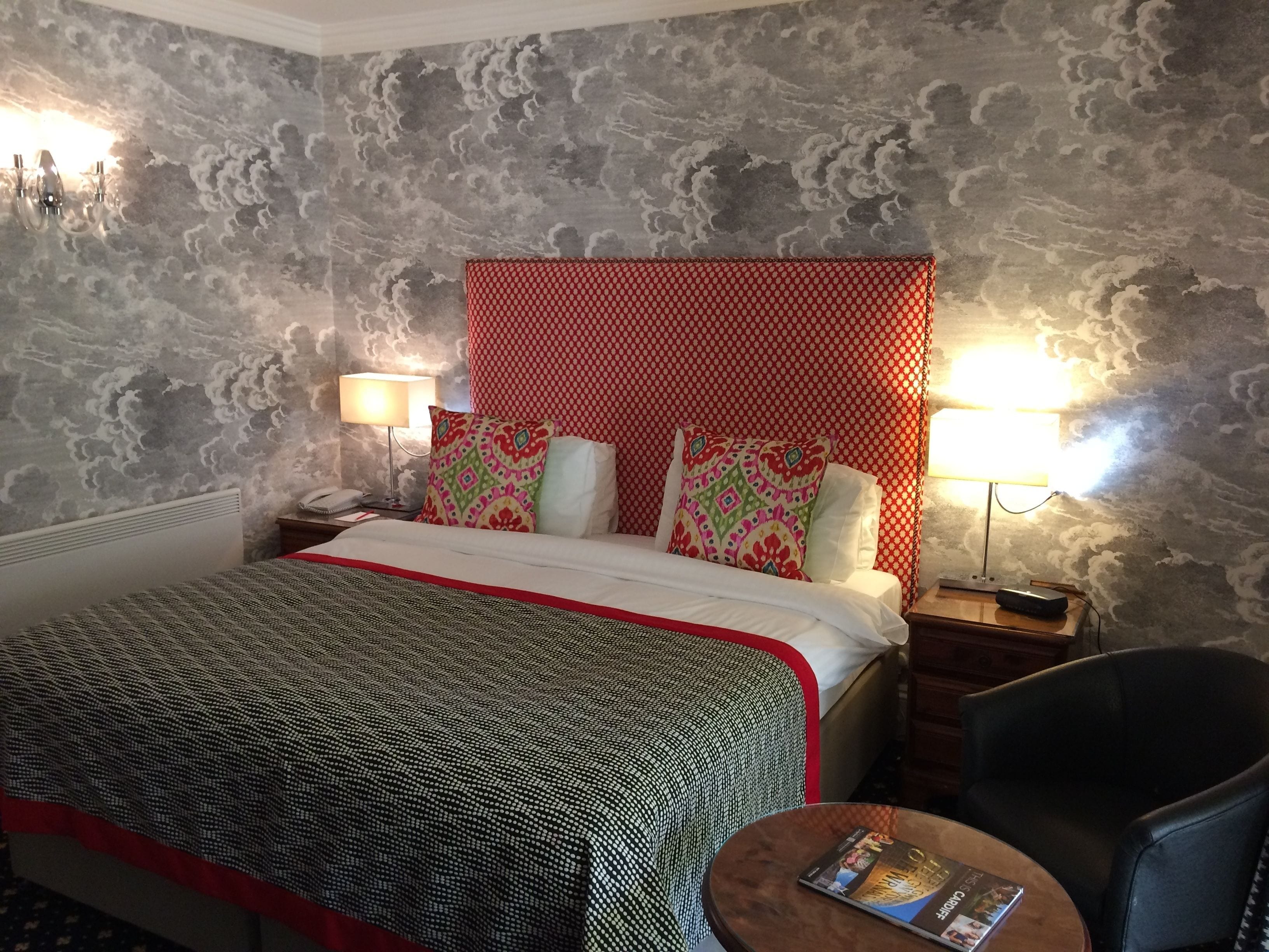 Habitación de hotel con cama, mesita de noche con lámpara, una silla y una pequeña mesa con una revista.