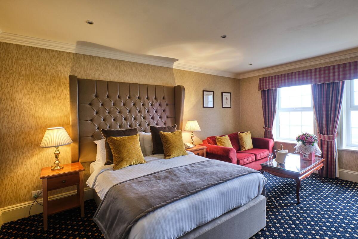 A hotel room with a large bed, a red sofa, a small table with a vase, and a window with curtains.