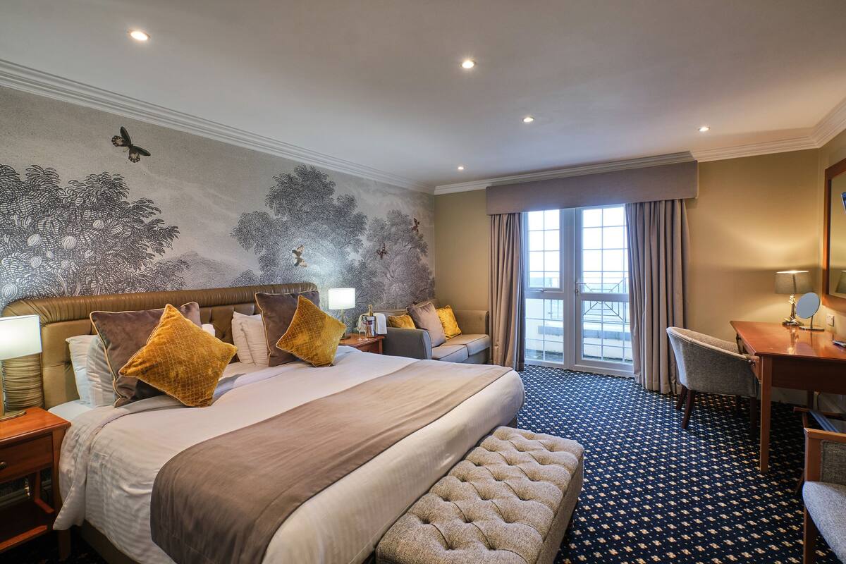A hotel room with a large bed, a desk, a chair, a bench, and a view of the outside through a window.