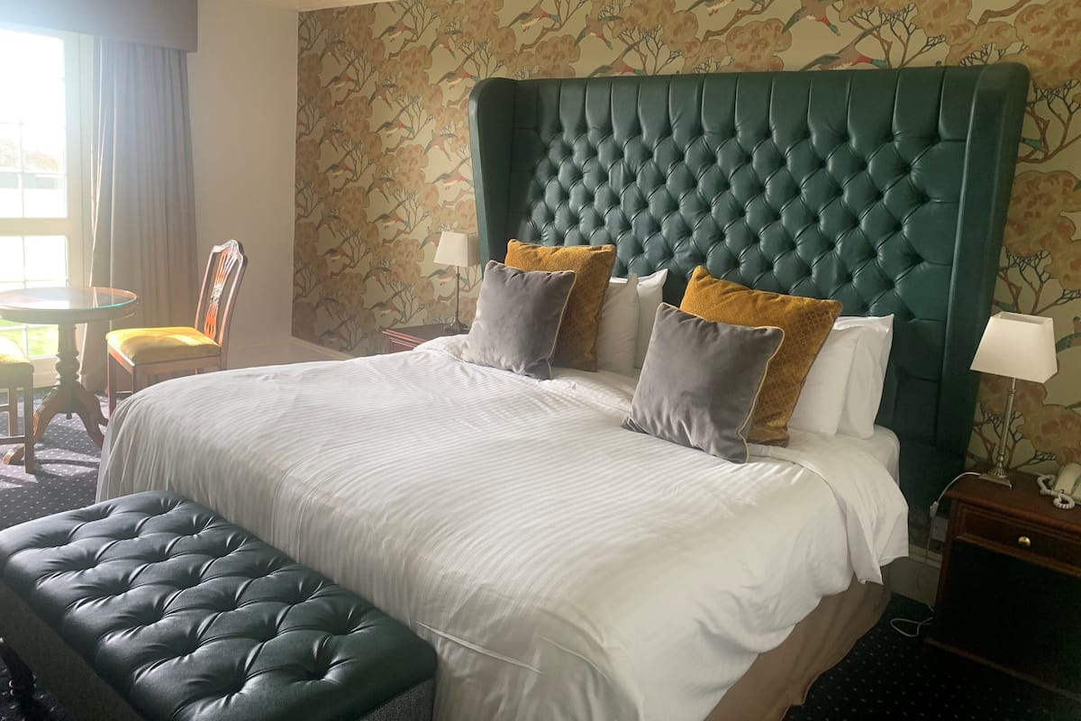 A hotel room with a large bed, a tufted headboard, a bench, a chair, and a small table.