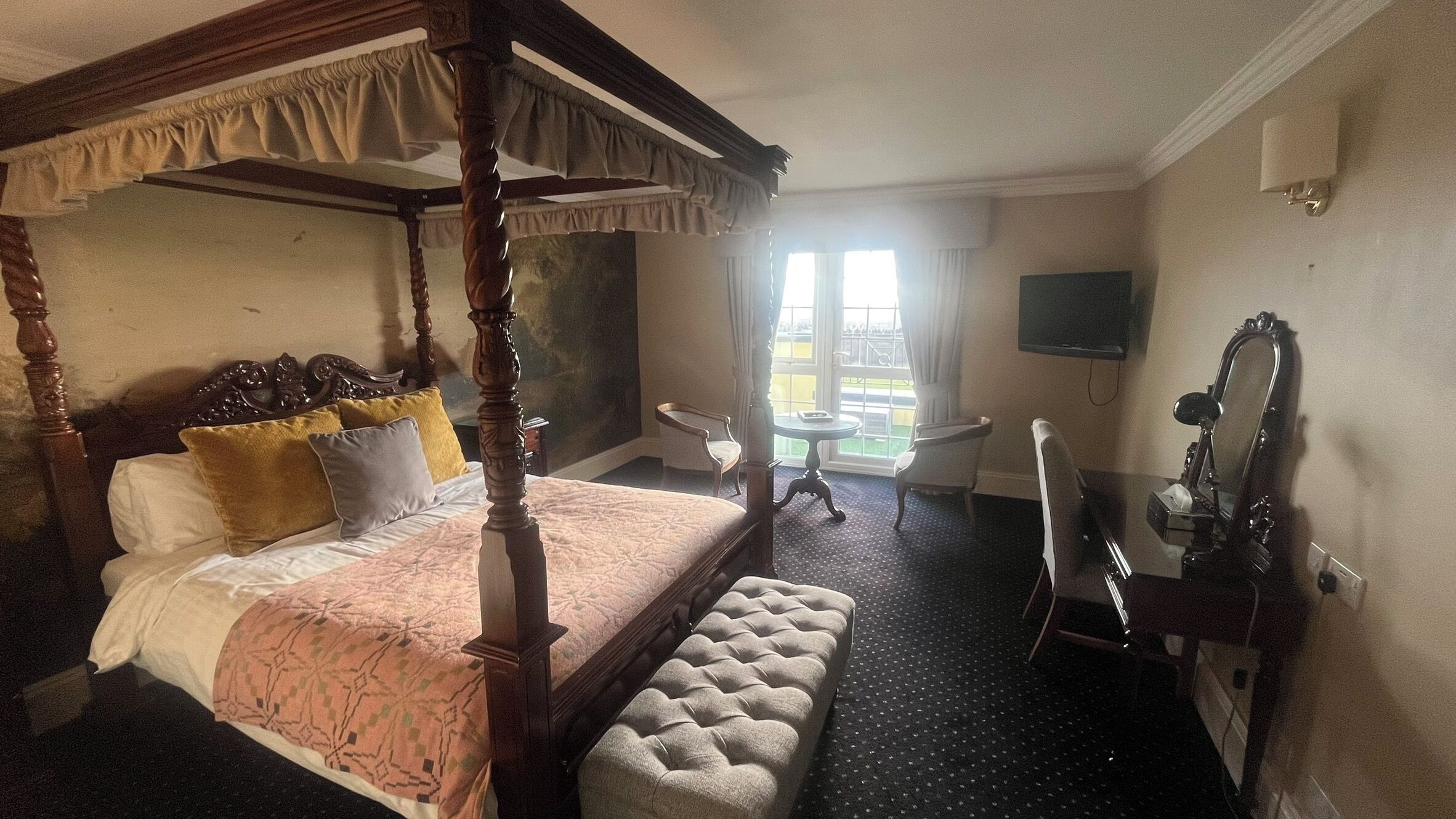 A four-poster bed with a bench, a desk, a chair, and a TV in a hotel room.