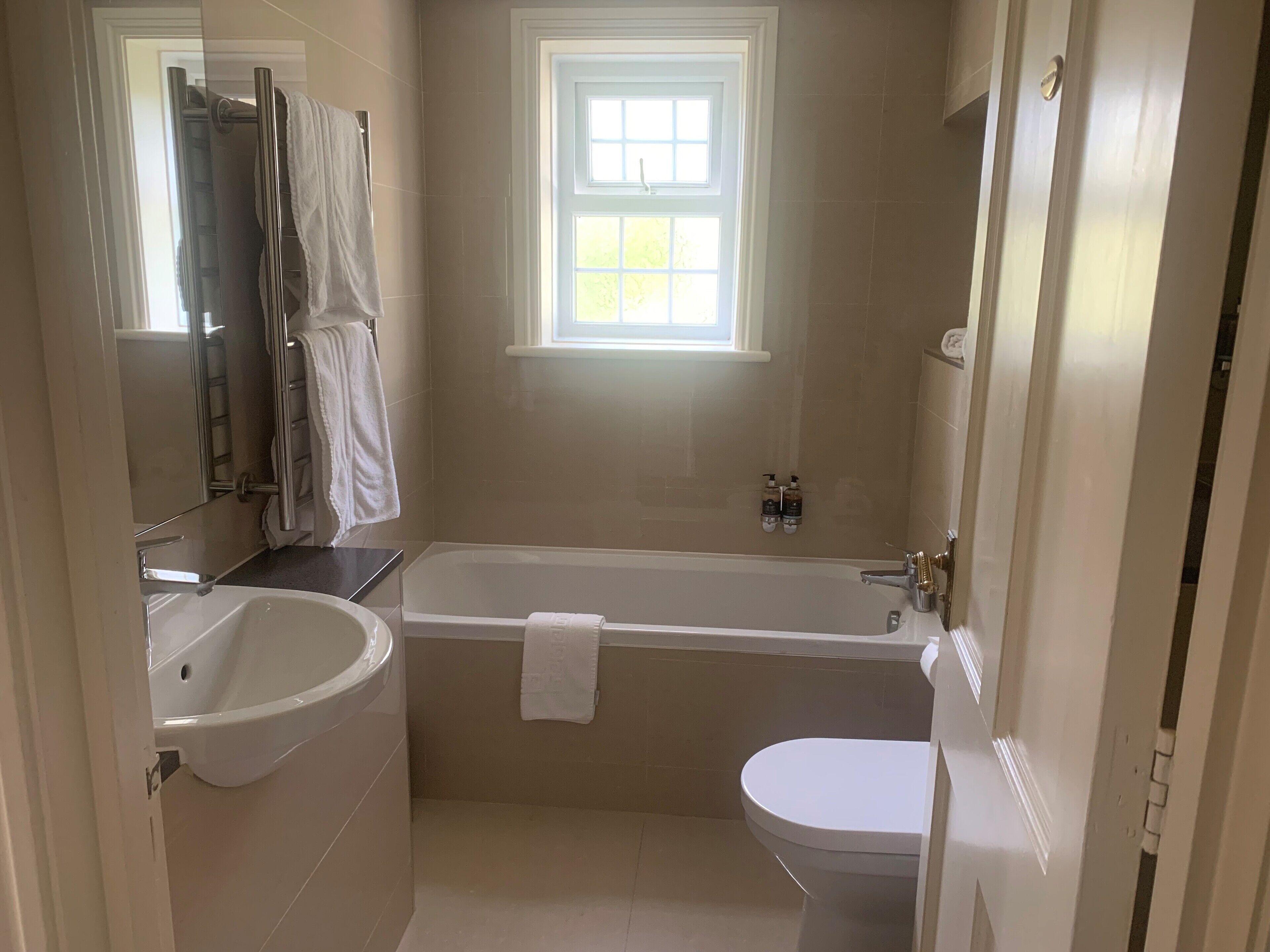 Baño con bañera, inodoro y lavamanos. Cuenta con una ventana que proporciona luz natural y un perchero con toallas.