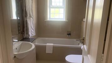 Baño con bañera, inodoro y lavamanos. Cuenta con una ventana que proporciona luz natural y un perchero con toallas.