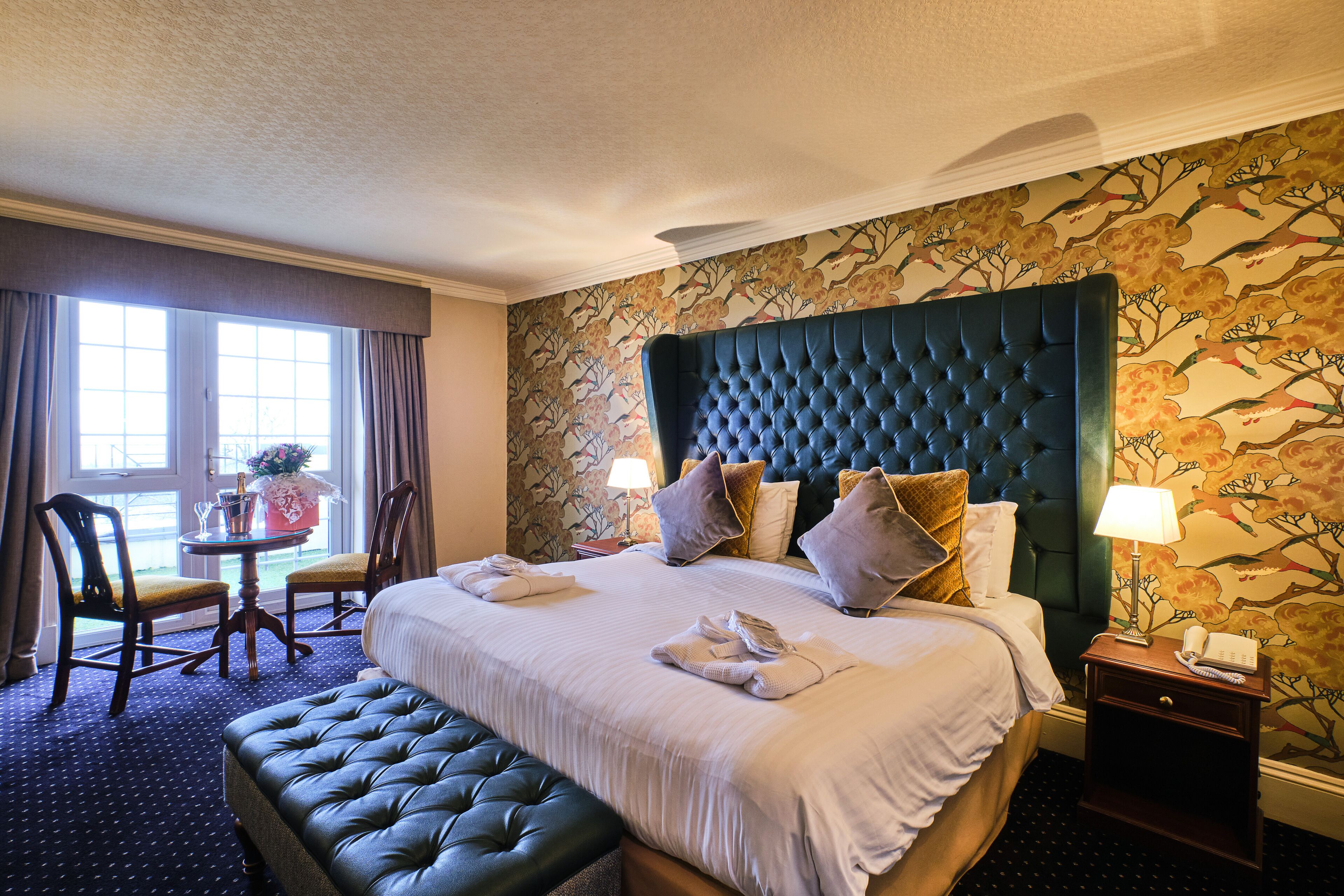 A hotel room with a large bed, a headboard, a bedside table with a lamp, a small table with a vase, and two chairs.
