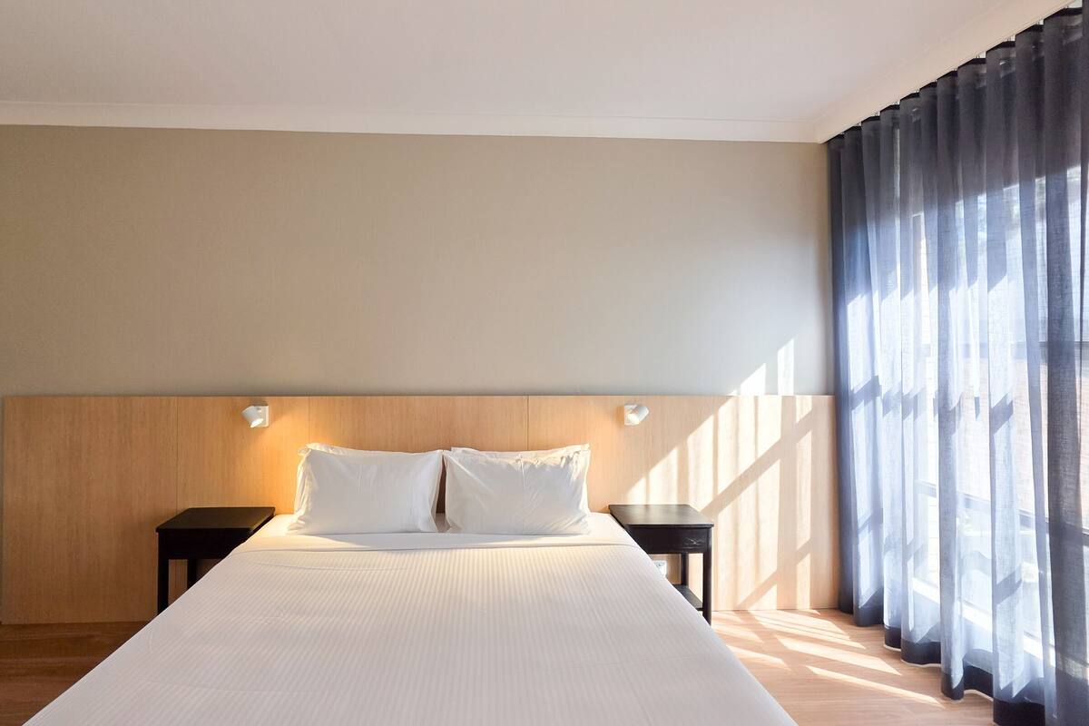 A hotel room with a large bed, two bedside tables, and a view through sheer curtains.