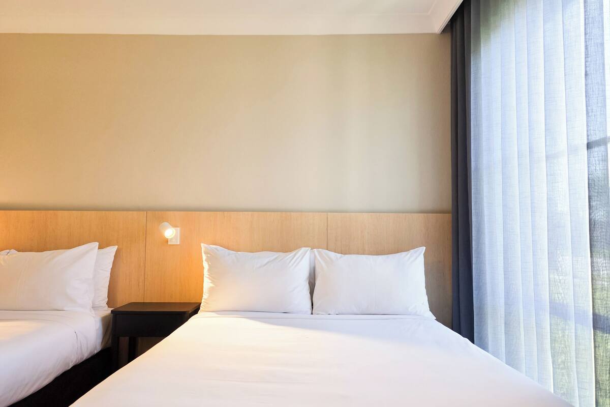 A hotel room with two beds, a wooden headboard, a nightstand with a lamp, and a window with sheer curtains.