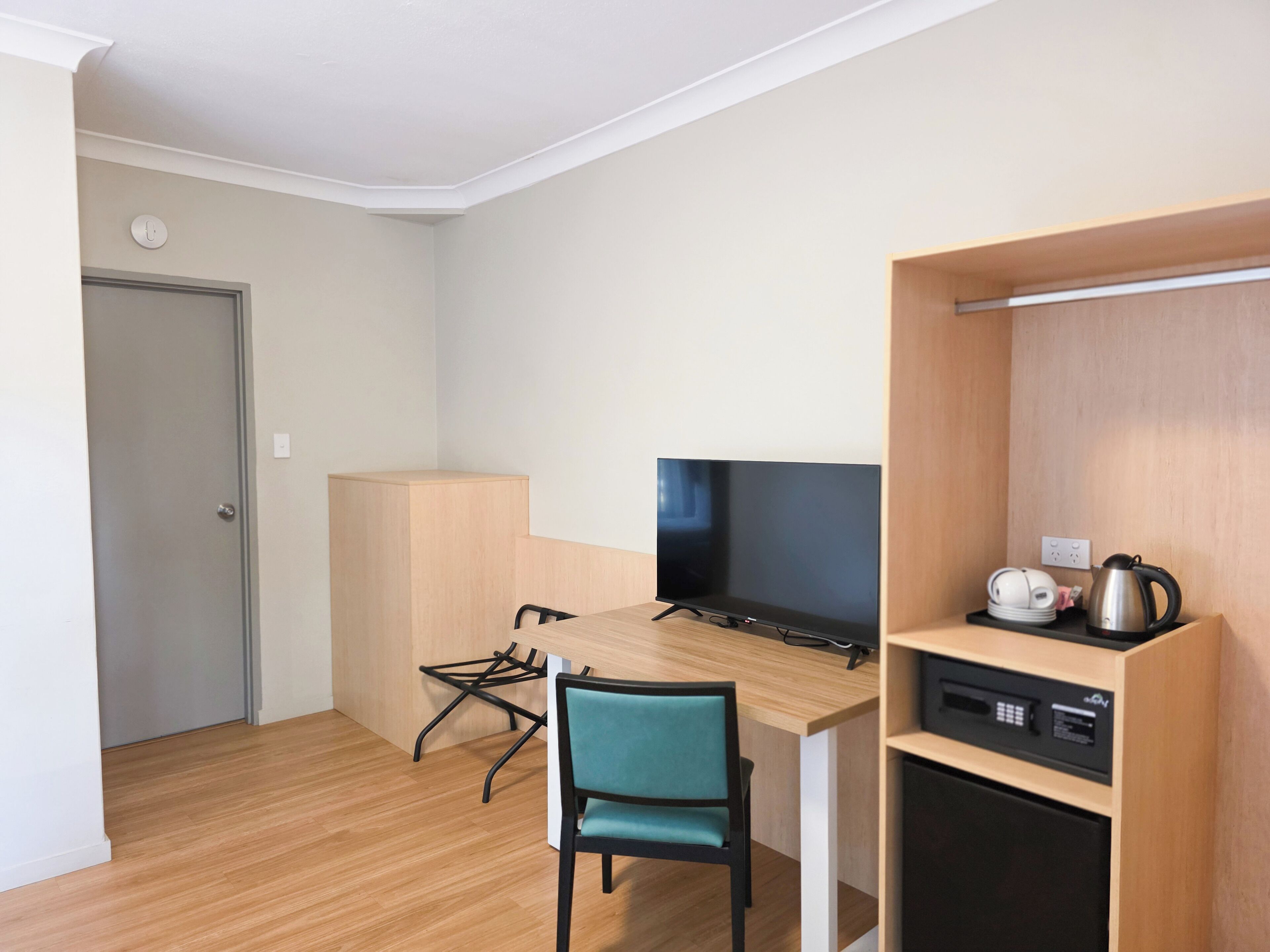 Quarto compacto com uma escrivaninha de madeira, televisão preta, cadeira verde-água, micro-ondas e chaleira em uma prateleira.